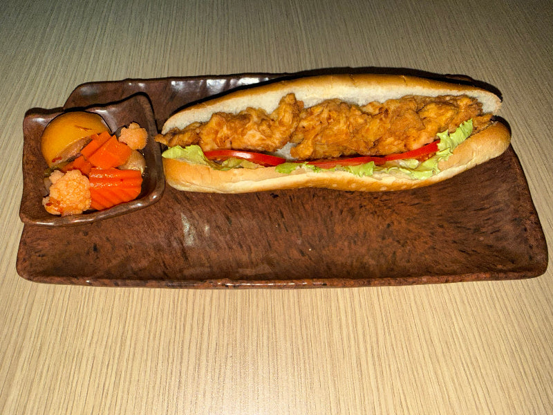 A fried chicken sub sandwich served on a dark brown ceramic rectangular plate with a small side dish of pickled vegetables, set on a light wood table.