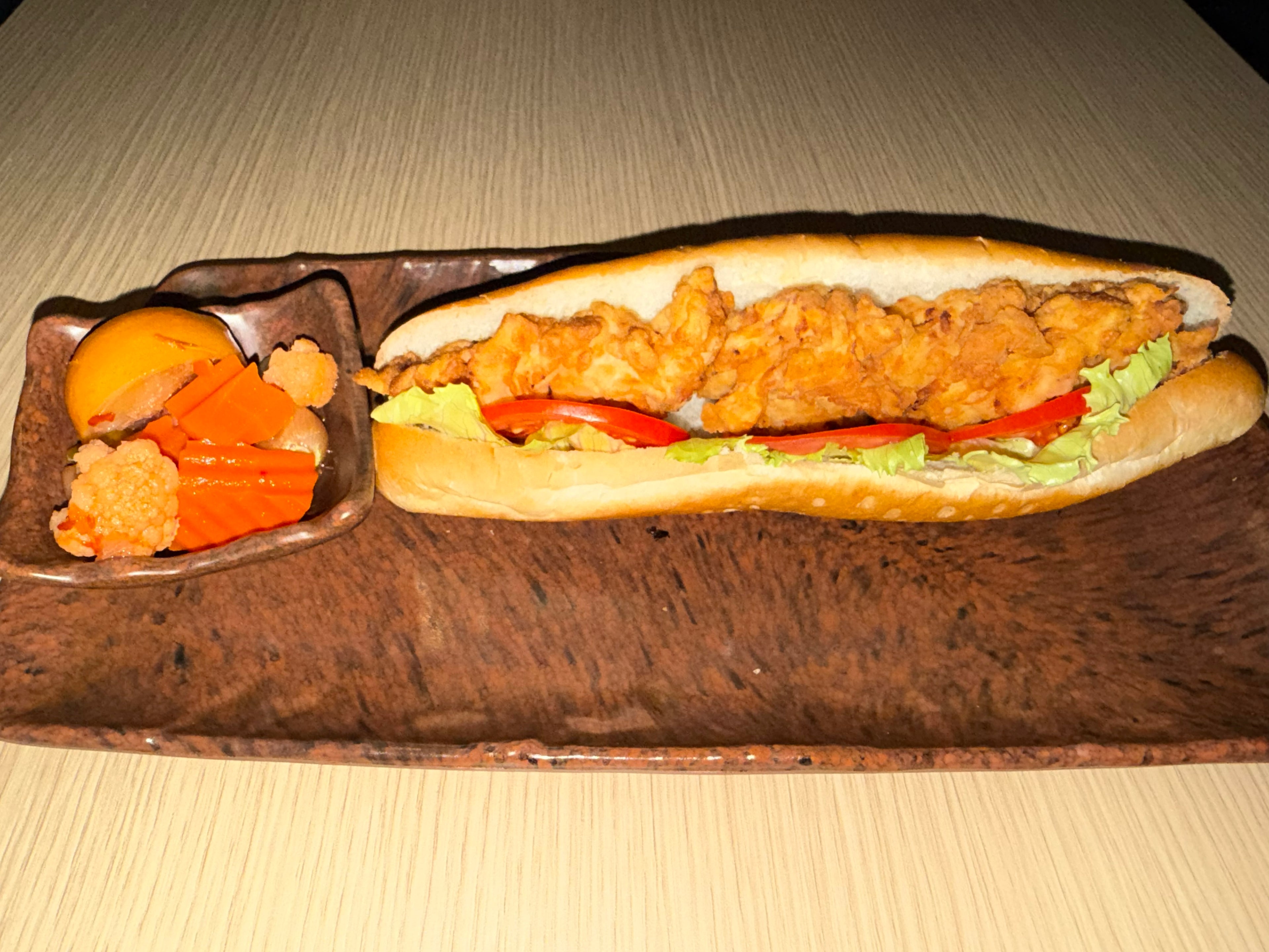 A fried chicken sandwich on a long wooden serving platter with a small side dish containing orange vegetables and a yellow item, set on a light wood table.