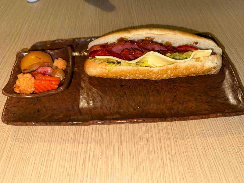 A gourmet hot dog served on a dark brown ceramic rectangular plate, accompanied by a small side dish of pickled vegetables and a yellow fruit, set on a light wood table.