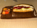 A gourmet hot dog served on a dark brown ceramic rectangular plate, accompanied by a small side dish of pickled vegetables and a yellow fruit, set on a light wood table.